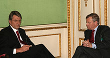 Двое пожилых мужчин в черных костюмах и красных галстуках сидят лицом друг к другу в комнате с зелеными, белыми и золотыми стенами.