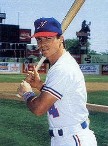 A man in a white baseball jersey with red and blue trim stands holding a bat