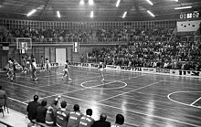 Photo en noir et blanc d'un match de basket. On distingue les remplaçants de Denain assis sur le banc au bord du terrain regardant les joueurs des deux équipes sous le panier.