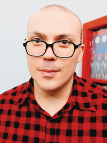 A hairless man with black plastic eyeglasses and a red flannel shirt in a room with a picture on the wall