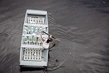 Overhead view of a young girl, alone, rowing through murky floodwaters on a wooden boat carrying several large plastic water bottles