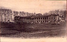 Photographie en noir et blanc imprimé en sépia sur carte postale. De jeunes gens jouent sur un terrain de rugby avec un bâtiment qui domine le terrain, derrière les poteaux de rugby.