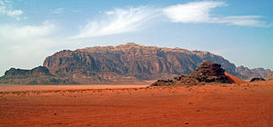 Širok skalnat rumen greben, ki sega do najvišje točke na sredini, nad puščavo rdečkastega peska z manjšo vzpetino v ospredju