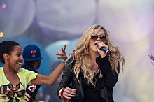 Kesha sur scène en noir, un microphone à la bouche, portant des lunettes accompagné d'autres personnes