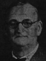A grainy image of a stern-faced, balding man with glasses.