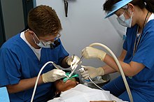 Dental assistant on the right supporting a dental operator on the left, during a procedure. US Navy 030620-N-8937A-002 Lt. William Peterson (left) of Branch Dental Clinic Sasebo, Japan drills a cavity while his dental assistant, Miho Otubo, ensures the area remains clean.jpg