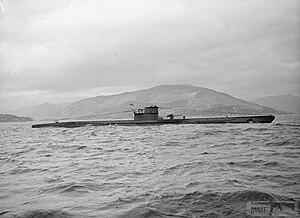 A side view of a surfaced submarine, seen in the middle distance; she is sailing in coastal waters and a mountainous landscape is in the background.