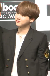 Suga, in a black suit, holds his hands behind his back, looking left