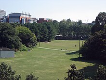 A grassy field surrounded by large, green-leafed trees and bushes.  A sidewalk cuts through the field horizontally. A large red brick and  white concrete building with idealized smoke stacks is in the background  to the left, as are a few other less visible buildings.