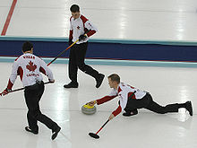 A imagem mostra três jogadores da equipe do Canadá. Um deles está na posição de lançamento (agachado) e os outros dois, em pé, se preparam para varrer o gelo.