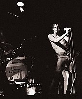 На сцене рок-группа. Слева - ударная установка. Певец Игги Поп поет в микрофон. Он в джинсах и без рубашки.