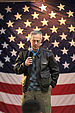 A color photo of an older white man in a leather jacket with a large American flag in the background. He is looking down toward the ground and is holding a microphone.
