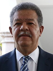 A portrait shot of a middle-aged man smiling somewhat and looking straight ahead. He has light brown skin, slightly African facial features, curly dark hair. He is mustachioed and wears a suit and tie.