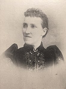 B&W portrait photo of a woman with her hair in an up-do wearing a dark, high-collared blouse.
