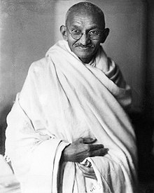 Wajah Gandhi di usia tua tersenyum, mengenakan kacamata, dan dengan sabuk putih di atas bahu kanannya