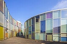 Farbfotografie von zwei modernen Gebäuden, die eine leicht kurvige Form haben. Zwischen ihnen verläuft ein Weg. Die Gebäude bestehen aus Glas mit bunten Jalousien und unterschiedlich großen Fenstern.