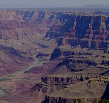 A grayish-colored river with some green vegetation on its banks but small compared to the high reddish and tan walls of the canyon it is in.