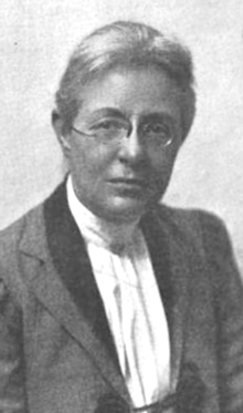 A middle-aged white woman with fair hair, wearing eyeglasses, a tailored jacket with lapels, and a white blouse with a pleated bib front and high collar