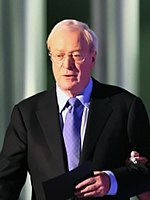 A picture of an old man wearing a black coat. He is wearing a light purple tie.