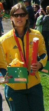 A standing blonde woman wearing sunglasses, a green and yellow track suit, and a bronze medal, while holding a plush kangaroo.