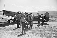 Groupe d'une dizaine d'hommes rassemblés autour d'un avion posé dans le désert.