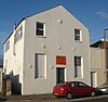 Three-quarter view of a tall, two-storey, plain-walled, white-painted building with a pointed roof. The front has one blank window and a half-blank, half-glazed window flanking a narrow door, above which is an orange sign. There are also six circular protrusions. The side wall has three windows and a thin metal flue.