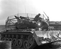 1953年3月25日、ドーザー付きのM46パットンの砲塔にバズーカ対策の金網を張っている海兵隊員。 戦争終盤のこの当時には国連軍戦車は壕に待機し砲塔だけ出して砲撃支援任務を行うことが主体となっており、共産軍に鹵獲されたバズーカによる攻撃を受けて大きな損害を出していた。