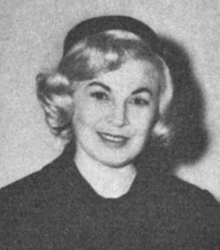 A smiling blond white woman wearing a dark coat and a dark pillbox hat