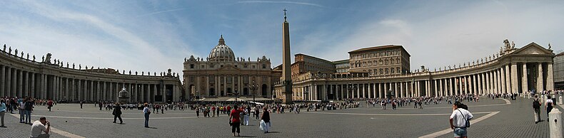 Panorama showing the facade of St. Peter's at the centre with the arms of Berninis colonnade sweeping out on either side. It is midday and tourists are walking and taking photographs.
