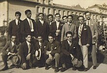 Photo en noir et blancs d'une dizaine de jeunes hommes qui posent en groupe. Ils portent des costumes, et on distingue un bâtiment de leur école à l'arrière-plan