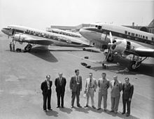 Image en plongée en noir et blanc. Sept hommes au premier plan, trois avions à l'arrière-plan.