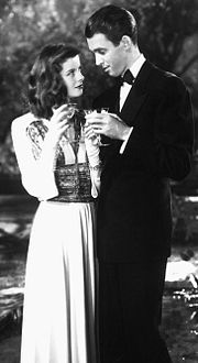 Hepburn and a smartly dressed man standing at night by a pool. She is holding a glass of champagne and they are looking at each other flirtatiously.