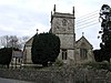 Grey stone building on 3 bays with a square stone tower at near end of central bay. To the left is a porch with slate roof. In front is a yew tree and gravestones behind a stone wall separating it from a road.