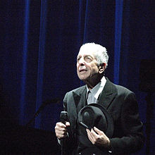 Muž držící v pravé ruce mikrofon a v levé klobouk