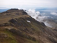 "A smattering of buildings situated on a flat-topped mountain with fog rolling off one side."