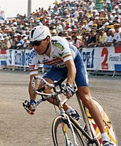 A man riding a bicycle wearing a helmet with a visor. Behind him there is a crowd of people on the road side