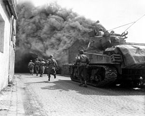 Infanteriști americani sprijiniți de un tanc M4 Sherman înaintează pe o stradă incendiată din Wernberg în aprilie 1945