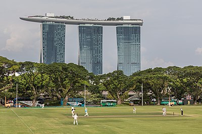 ’n Krieketwedstryd met spelers van die krieketklub in Padangpark met die Marina Bays Sands Hotel op die agtergrond, soos gesien van die Singapoerse Nasionale Galery.