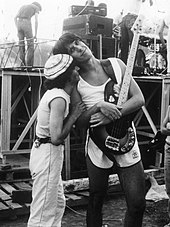 A black and white image of a man in a white outfit and a white hat hugging a taller man wearing a white t-shirt and shorts holding an electric guitar