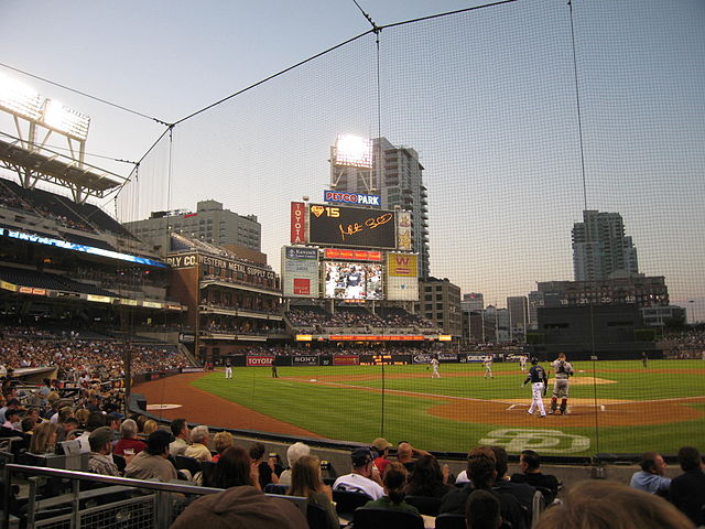Half of a baseball stadium at dusk, seen from behind home plate during a game, with tiers rising high on the left. Curving away from the camera from right to left is a sheet of black netting.