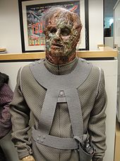 A mannequin wearing a face mask that resembles decomposing flesh and a grey body suit.