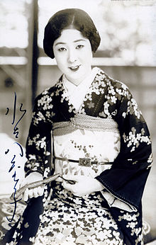 A young Japanese woman wearing a print kimono, smiling, seated, holding an object in her hands