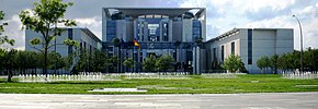 Frontale Farbfotografie vom Bundeskanzleramt mit Rasen und vielen kleinen Wasserfontänen. Vor dem Eingang stehen eine Europaflagge und eine Deutschlandflagge. Das Hauptgebäude besteht aus viel Glas und einigen Betonstützen und wird von zwei quaderförmigen Seitenflügeln flankiert.