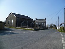 plain tarmac road passing to the right of a large dark-rendered chapel with two-storey house to the side