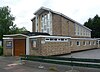 Three-quarter view of a pale brick building with prominent white soffits. The entrance area in the left foreground is flat-roofed and has a wide brown door flanked by posters. Above this, a tall window reaches to the slightly sloping roofline; it is split into nine unequal panes. There are six windows of decreasing height on the side elevation.