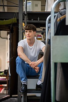 Ein junger weißer Mann mit braunen Haaren sitzt auf einer Metalltreppe, er trägt blaue Jeans und ein weißes T-Shirt.