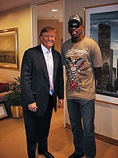 Donald Trump posing with former basketball player Dennis Rodman in a room with paintings adorning the walls. Trump is wearing a suit with a light-colored tie and dress shirt, while Rodman is wearing a brown t-shirt with a design on it, blue jeans, and a baseball cap that also has a design on it.