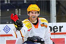 Photographie couleur d’un hockeyeur, de buste, portant un protège-dents multicolore et un maillot blanc avec un ours noir