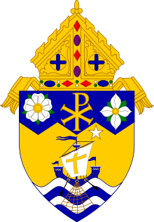 Shield topped by a mitre, featuring a white heraldic rose, Chi Rho, and Pacific dogwood on blue field at top; the Barque of St. Peter and a white star on gold field; and four alternating blue and white wavy stripes at the bottom