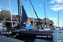 Voilier noir amarré au ponton. On voit le foil tribord, qui est vert. En arrière-plan, la base de sous-marins allemande de Lorient.